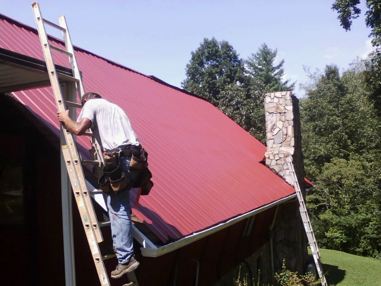 Roofer on a ladder beside a red metal roof and stone chimney in Pikeville